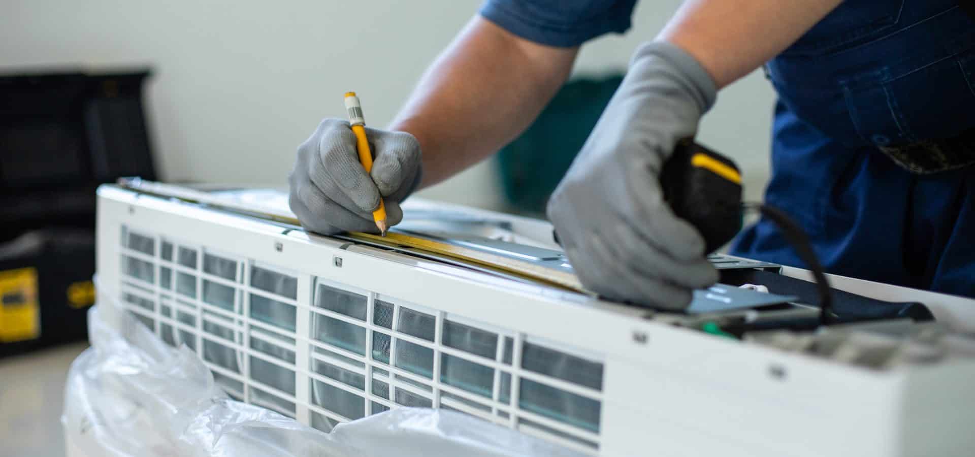 Male technician makes mark with pencil when measuring air conditioner unit. Male technician makes mark with pencil when measuring air conditioner unit. Close up of man's hand in protective gloves holds pencil and puts marks measuring air conditioner with tape measure.